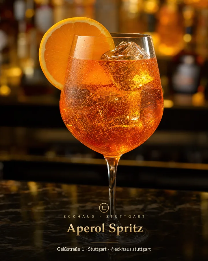 Aperol Spritz im Weinglas mit Eis und Orangenscheibe auf der Theke, dahinter die beleuchtete Flaschenwand der Bar.